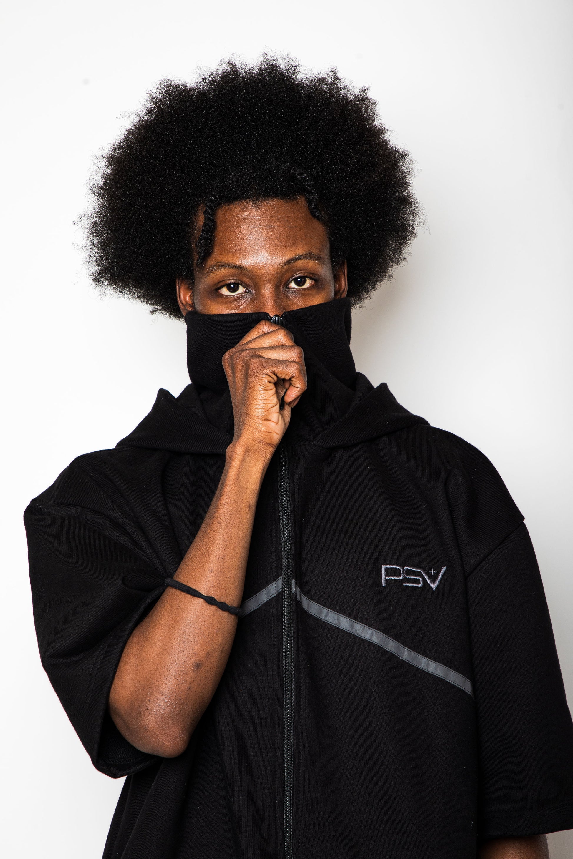 Person wearing a black hoodie with a logo, covering their face with one hand on a white background